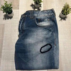 Blå jeans från Philipp Plein - Säljer ett par blå jeans från Philipp Plein med snyggt slitna detaljer och klassisk femficksdesign. Jeansen har normal passform och raka ben, med läderpatch bak i midjan. Perfekta för dig som gillar en avslappnad men ändå stilren look. Stolek 33!