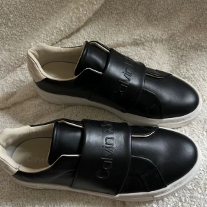Calvin Klein slip ons - Hejsan, gör ett försök att sälja mina oanvända skor. Beställde dessa från zalando men var för lat för att göra retur så det har bara stått i min hall. Skorna är super snygga men tog helt enkelt en för liten storlek men inbillade mig själv att de kanske skulle passa med tiden vilket dom ej gjorde 🤣.  Säljer alltså pga liten strl .  Storlek 41. Färg svarta med beige detalj bak. Vit sula. Inköpspris 1495 (zalando) 