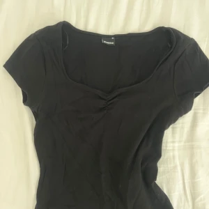 Svart topp med rynkad detalj - Svart kortärmad topp från gina tricot med snygg rynkad detalj vid bysten och djupare rundad urringning. Toppens passform är figurnära och materialet är mjukt och stretchigt, perfekt för en stilren look.