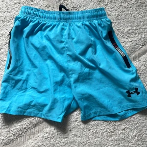 Blå träningsshorts från Under Armour - Snygga ljusblå träningsshorts från Under Armour med elastisk midja och svart logga broderad på benet. Shortsen har dragkedjeförsedda fickor och är gjorda i lätt polyester som andas. Perfekta för gymmet eller löprundan.