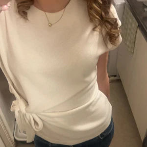 Topp med knytning - Säljer en stilren vit topp med knytdetalj i midjan. Toppen har korta ärmar och är tillverkad i ett mjukt material som ger en snygg passform. Perfekt att matcha med jeans eller kjol.
