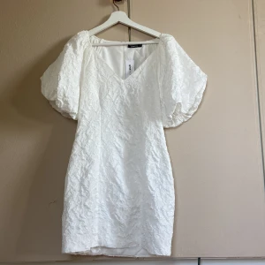 Vit brokadklänning med puffärmar - Superfin vit brokadklänning med strukturmönster och v-ringning. Klänningen har kort längd och stora puffärmar som ger en trendig siluett. Perfekt för somriga tillfällen eller studentfirande. Stängs med dragkedja i ryggen.