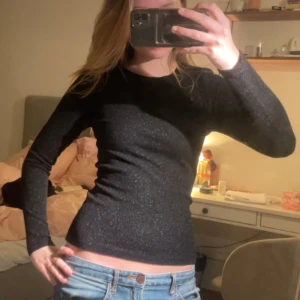 Svart glittrig långärmad topp från Mads Nørgaard - Säljer en svart långärmad topp från Mads Nørgaard med glittrigt material på utsidan och mjukt typ på insidan. Sitter tight och har en enkel, stilren design som passar perfekt till jeans eller kjol. Superfin att styla till olika tillfällen!