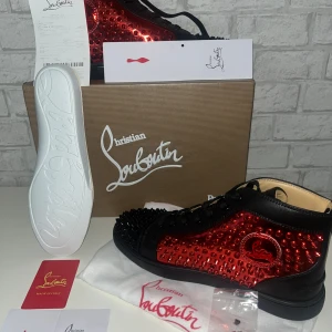 Christian Louboutin sneakers med nitar - Säljer ett par Christian Louboutin sneakers i svart och röd med glansig finish och svarta nitar över hela skon. Skorna har snörning, rund tå och platt sula. Ikonisk Louboutin-logga på sidan och vit sula undertill. (20)