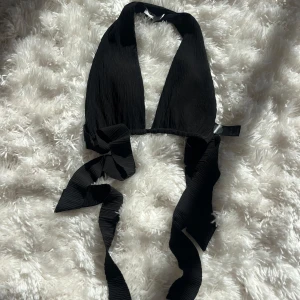 Svart halterneck topp med knyt från NA-KD - Säljer en svart halterneck topp från NA-KD med breda knytband i midjan och djup v-ringning. Toppen är ärmlös och har en croppad passform, perfekt för att skapa en trendig look.