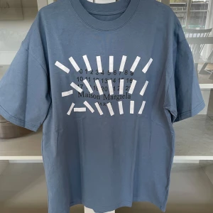Blå t-shirt från Maison Margiela - Säljer en blå t-shirt från Maison Margiela med ikoniskt tryck framtill i vitt och svart. T-shirten har rund halsringning och korta ärmar. Tillverkad i Italien och i mjuk bomull. Perfekt för dig som gillar stilrena designer med en twist.