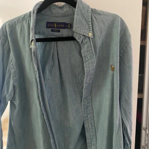 Ljusblå skjorta från Ralph Lauren - Säljer en ljusblå skjorta från Ralph Lauren i slim fit-modell. Skjortan har klassisk krage, knappar framtill och den ikoniska broderade loggan på bröstet. Perfekt för dig som gillar stilrena och tidlösa plagg.
