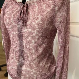 Mönstrad offshoulder långärmad tröja - Säljer en tunn och luftig blus med lång ärm och paisleymönster i rosa och vitt. Blusen har knytning vid halsen och en avslappnad passform. Perfekt för dig som gillar boho-stil!