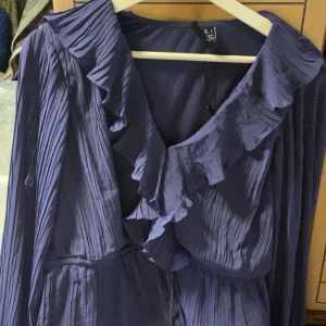 Mörkblå plisserad klänning med volang - Säljer en mörkblå plisserad klänning från Vero Moda med v-ringning och volangdetaljer längs halsringningen. Klänningen har långa ärmar och ett elegant fall, perfekt för dig som gillar en feminin stil.