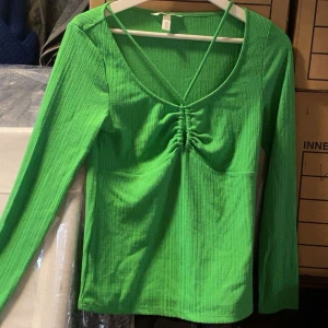 Grön långärmad topp med rynkdetalj - Säljer en grön långärmad topp med djup v-ringning och rynkdetalj framtill. Toppen har smala band som korsas i ryggen och är tillverkad i ett ribbat material. Perfekt för dig som vill sticka ut med färg!