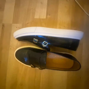 Svarta loafers med spännen och vit sula - Säljer ett par svarta loafers med två dekorativa metallspännen på ovansidan och en tjock vit platt sula. Skorna har en stilren design och är tillverkade i ett material som liknar skinn. Perfekta för dig som vill ha en snygg och bekväm sko.
