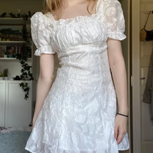 Vit spets klänning med puffärmar - Vit klänning med puffiga kortärmar och ett broderat blommönster. Klänningen har en söt, rynkad detalj vid halsringningen och en kort längd. Finns dragkedja på baksidan. Perfekt till student! HELT OANVÄND