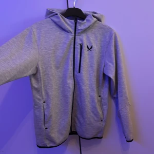 Grå huvjacka från SOC - Säljer en grå huvjacka från SOC med svarta dragkedjor och logga på bröstet. Jackan har huva, två sidofickor och en bröstficka med dragkedja. Perfekt för en sportig stil.