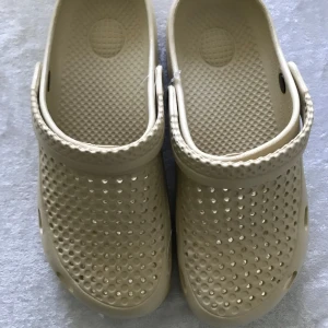 Beige Tofflor  - Säljer ett par beige tofflor med klassisk design och perforerad ovandel. Skorna har en justerbar hälrem och platt sula, perfekta för sommardagar eller som inneskor.