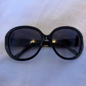 Svarta solglasögon från Jimmy Choo med glittriga skalmar - Snygga svarta vintage solglasögon från Jimmy Choo med stora, ovala bågar och glittriga detaljer på skalmarna. Märket syns  på sidan. Perfekta solglasögonen för sommaren. Kommer i Prada fodral då jag tappa bort originalfodralet. Skriv om ni har frågor eller vill ha fler bilder💗
