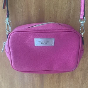 Rosa axelväska från Victoria's Secret - Säljer en snygg rosa axelväska från Victoria's Secret med silverfärgad logga framtill. Väskan har justerbar axelrem och dragkedja upptill. Perfekt för att bära det viktigaste när du är på språng.
