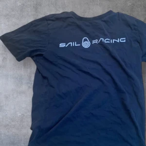 Svart t-shirt från Sail Racing - Svart t-shirt från Sail Racing med tryckt logga på ryggen och liten logga i nacken. Klassisk passform och rund halsringning. Perfekt för dig som gillar stilrena plagg med sportig känsla. Storlek 160cm. Den har ett litet hål som man knappt ser på framsidan.
