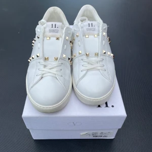 Vita Valentino Garavani sneakers med nitar - Säljer ett par vita Valentino Garavani sneakers med ikoniska guld- och silverfärgade nitar. Skorna har rund tå, platt sula och är tillverkade i skinn. Klassisk och stilren design med snörning och detaljerade nitar längs sidorna. Hör av dig vid frågor 