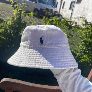 Vit bucket hat från Polo Ralph Lauren - Vit bucket hat från Polo Ralph Lauren med klassisk blå broderad logga framtill. Hatten har bättre bred och är tillverkad i bomull, perfekt för soliga dagar.