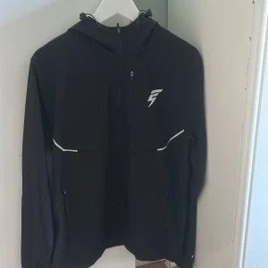 Svart vindjacka från Elude med reflexdetaljer - Svart track jacket från Elude Jackan har reflexdetaljer på bröstet, ärmarna och ryggen samt en logotyp på bröstet och huvan. Storlek M , pris kan diskuteras , (aldrig använd)