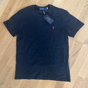 Svart ralph lauren tshirt - Säljer en klassisk svart t-shirt från Polo Ralph Lauren med rund hals och kort ärm. T-shirten har den ikoniska röda broderade loggan på bröstet. Perfekt basplagg med stilren design. Skit fet nu till sommarn. Står storlek L men det är barnstorlek så det motsvarar vanlig S. Den är helt ny med etiketten på!