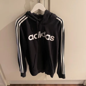 Svart hoodie från Adidas med vita ränder - Svart hoodie från Adidas med klassiska vita ränder längs ärmarna och stor vit logga på bröstet. Tröjan har huva med dragsko och en stor magficka framtill. Perfekt för en sportig och avslappnad stil.