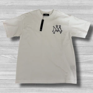 Amiri T-shirt - Säljer en vit t-shirt från Amiri med svart tryck på bröstet och stor AMIRI-logga på ryggen. Klassisk passform och rund halsringning. Perfekt för dig som gillar stilrena och exklusiva plagg.