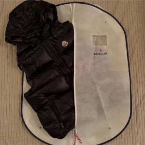 Svart dunväst från Moncler med huva - Säljer en svart dunväst från Moncler med avtagbar huva och dragkedja framtill. Västen har två fickor med dragkedja och klassisk Moncler-logga på bröstet. Insidan har ett unikt Moncler-tryck och etikett. Man kan även scana. 😇