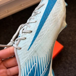 Nike fotbollsskor i ljusblått och blått - Säljer ett par Nike fotbollsskor i ljusblått med blå detaljer och stor Nike-logga på sidan. Skorna har snörning och dobbar för spel på gräs. Snygg och sportig design, perfekt för dig som vill sticka ut på planen.