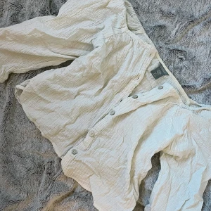 Vit skjorta från Lindex - Säljer en vit skjorta från Lindex i krinklad bomull med knappar framtill. Skjortan har halv arm och en avslappnad passform, perfekt för en stilren look. Klassisk krage och mjukt material.