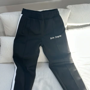 Svarta träningsbyxor från Palm Angels med vita ränder - Säljer ett par svarta träningsbyxor från Palm Angels med vita dubbla ränder längs sidorna. Byxorna har resår i midjan och fickor framtill. Märkeslogga broderad på ena benet. Perfekta för en sportig och trendig look. OBS!! Inte äkta. 