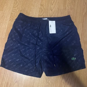 Mörkblå badshorts från Lacoste - Snygga mörkblå badshorts från Lacoste i helt nytt skick i stolek L. Men skulle säga de passar M mycket bättre. Den är lite liten i storlek. Den är helt oanvänd med alla etiketter kvar. Vid fler frågor är det bara att höra av sig!