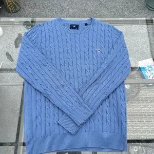 gant kabelstickad tröja herr blå bomull stl L - Klassisk och tidlös herrtröja från Gant i kabelstickad bomull – en riktig garderobshjälte! Modellen är i en mjuk och lite tjockare bomullskvalitet med rund hals och broderad logo. Färgen är en fräsch ljusblå som passar perfekt till både vardags och mer dressade outfits.  Storlek L, normal i passform. Inga defekter – aldrig använd! Perfekt för dig som gillar preppy stil, clean aesthetic eller bara letar efter ett bra basplagg i hög kvalitet.  Nypris runt 1300 kr. (PRISET DISKUTERBART)
