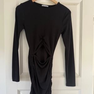 Svart långärmad klänning med rynkade detaljer - Säljer en svart långärmad klänning med rund hals och snygga rynkade detaljer framtill samt sidan. Klänningen har en tight passform och är perfekt för dig som gillar stilrena plagg med en twist. Öppen vid magen.