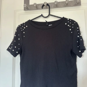 Svart ribbad topp med pärlor - Säljer en svart ribbad topp med korta ärmar och dekorativa pärlor på axlar och ärmar. Toppen har en rund halsringning och är stretchig i materialet. Perfekt för att lyfta en enkel outfit!