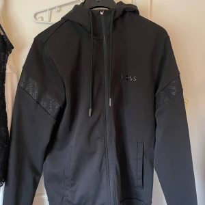 HUGO BOSS hoddie - Svart hoodiejacka från HUGO BOSS med dragkedja och luva. Jackan har diskreta BOSS-loggor på ärmarna och en stilren logotyp på bröstet. Storlek S! Tröjan är äkta går att scanna!