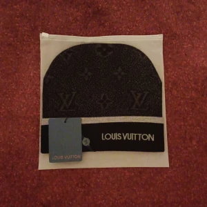 Svart mössa från ett välkänt märke  - Exklusiv svart mössa från ett välkänt märke med diskret monogrammönster och vit rand. Märkets logotyp syns tydligt framtill. Mössan är tillverkad i ett mjukt material och har en klassisk passform.