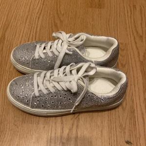 Glittriga sneakers med vita snören - Säljer ett par silvriga sneakers täckta med glittriga paljetter och vita snören. Skorna har en tjock vit sula och rund tå, perfekt för dig som vill sticka ut med din stil.