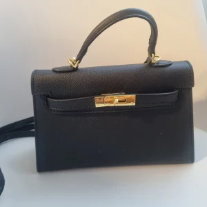 Supersöt handväska - Snygg svart axelväska i "skinnliknande" material med guldfärgat spänne och handtag upptill.Väskan har en stilren och klassisk design med både axelrem och handtag, perfekt för att lyfta din outfit. Ca 22 cm bred och 14 cm hög.