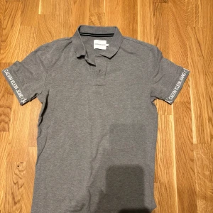 Grå pikétröja från Calvin Klein Jeans - Säljer en stilren grå pikétröja från Calvin Klein Jeans. Tröjan har klassisk krage och korta ärmar med logotypen tryckt på båda ärmarna. Perfekt för en clean och avslappnad look.