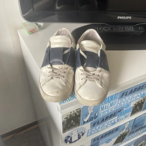 Valentino Garavani Open sneakers i vitt och marinblått - Säljer ett par snygga Valentino Garavani Open sneakers i vitt skinn med en bred marinblå detalj över sidan. Skorna har vita snören, rund tå och platt sula med ikoniska nitar baktill. Perfekta för dig som gillar stilrena och exklusiva sneakers.