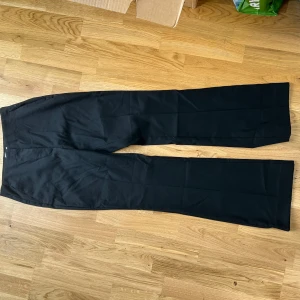 Svarta kostymbyxor från H&M - Klassiska svarta kostymbyxor från H&M med rak passform / lite bootcut och diskreta bakfickor. Byxorna har knapp och dragkedja i midjan och är perfekta för en stilren look. Materialet känns mjukt och har en lätt stretch.