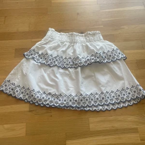 Vit kjol med broderade blommor från LMTD - Supersöt vit kjol från LMTD med broderade blommor i blått längs volangkanten och nederkanten. Kjolen har resår i midjan och dubbla volanger för extra volym. Perfekt för en fräsch och somrig look!