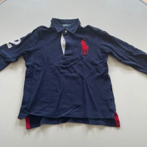  pikétröja från Polo Ralph Lauren - Mörkblå långärmad pikétröja från Polo Ralph Lauren med stor röd logga på bröstet och vit detalj vid knäppningen. Tröjan har krage och nummerdetalj på ena ärmen. Perfekt för en sportig och klassisk stil. För barn 