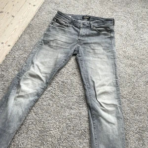 Grå slim fit jeans från Jack & Jones - Snygga grå jeans från Jack & Jones i modellen Slim/Glenn. Jeansen har klassisk femficksdesign, dragkedjegylf och en tvättad look med ljusa partier. Perfekta för dig som gillar en smal passform och stilren vardagsstil.