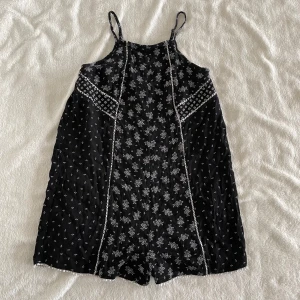 Svart mönstrad kort playsuit från Bershka - Säljer en svart kort playsuit från Bershka med vita blommiga och geometriska mönster. Den har smala axelband och är ärmlös, med dekorativa sömmar framtill. Perfekt för varma dagar!
