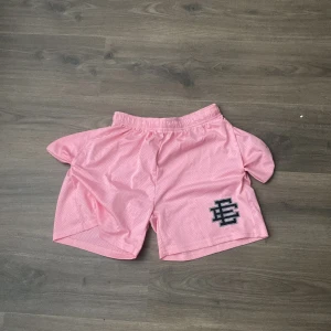 Eric Emanuel Shorts - Säljer ett par rosa meshshorts från Eric Emanuel med svart logga på vänster ben. Shortsen har elastisk midja och snörning för justerbar passform. Perfekta för träning eller avslappnade dagar. Storlek M.