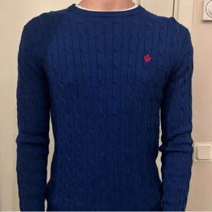 Blå kabelstickad tröja från Morris - Säljer en blå kabelstickad tröja från Morris i 100% merinoull. Tröjan har rund halsringning och en liten röd broderad logga på bröstet. Perfekt för dig som gillar klassisk stil med en modern touch.