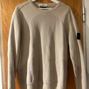 Beige sweatshirt från Stone Island - Säljer en stilren beige sweatshirt från Stone Island med klassisk rund halsringning och ribbade muddar. Tröjan har den ikoniska Stone Island-loggan på vänster ärm. Perfekt för en clean och avslappnad look. Låda samt kvitto finns även :)