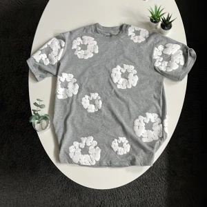 Grå t-shirt från Denim Tears - Säljer en grå t-shirt från Denim Tears med stora vita blommor tryckta över hela tröjan. T-shirten har rund hals och korta ärmar. Perfekt för dig som gillar streetwear och unika prints.
